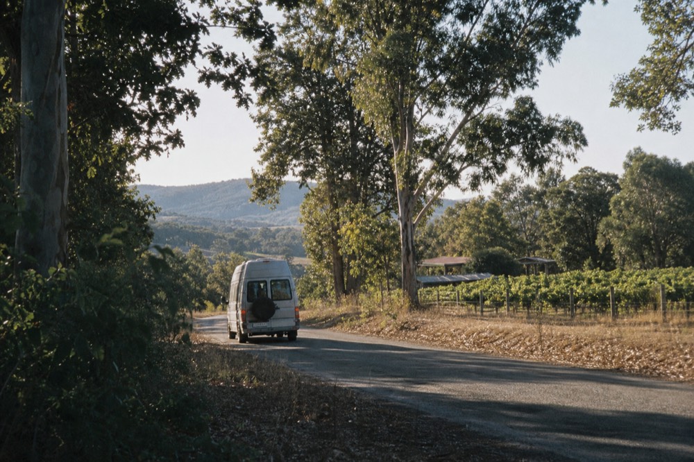 A small touring van on a quiet valley road between vineyards toward distant hills, morning light, no text, no logos, no faces