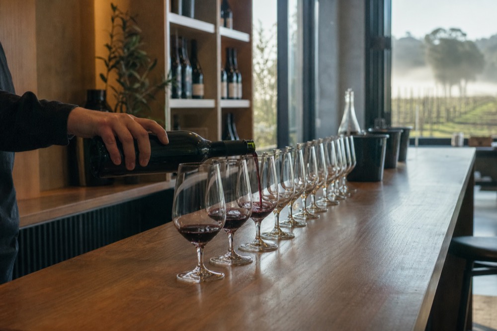 A wine host pouring into a row of tasting glasses on a wooden counter with bottles behind, warm interior light, no text, no logos, no people