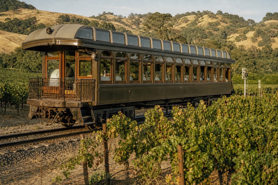 A restored early-1900s dining rail car passing between vineyard rows in Napa Valley, white-linen tables and wine glasses at the windows, golden hills beyond