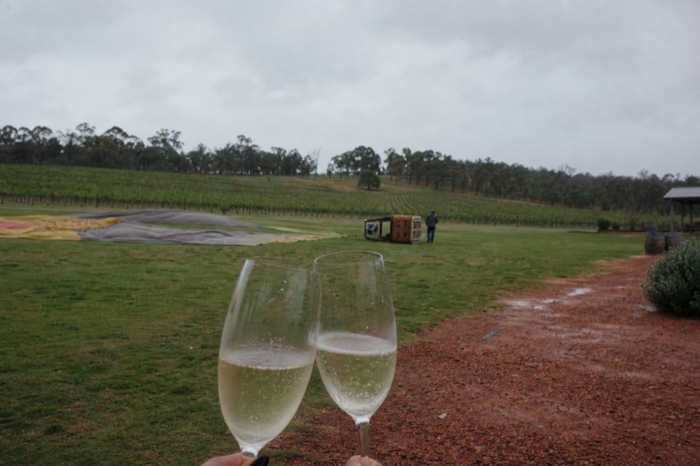 Two glasses of sparkling wine held up in a grassy field with a deflating balloon and vineyards behind after a morning flight, no text, no logos, no people