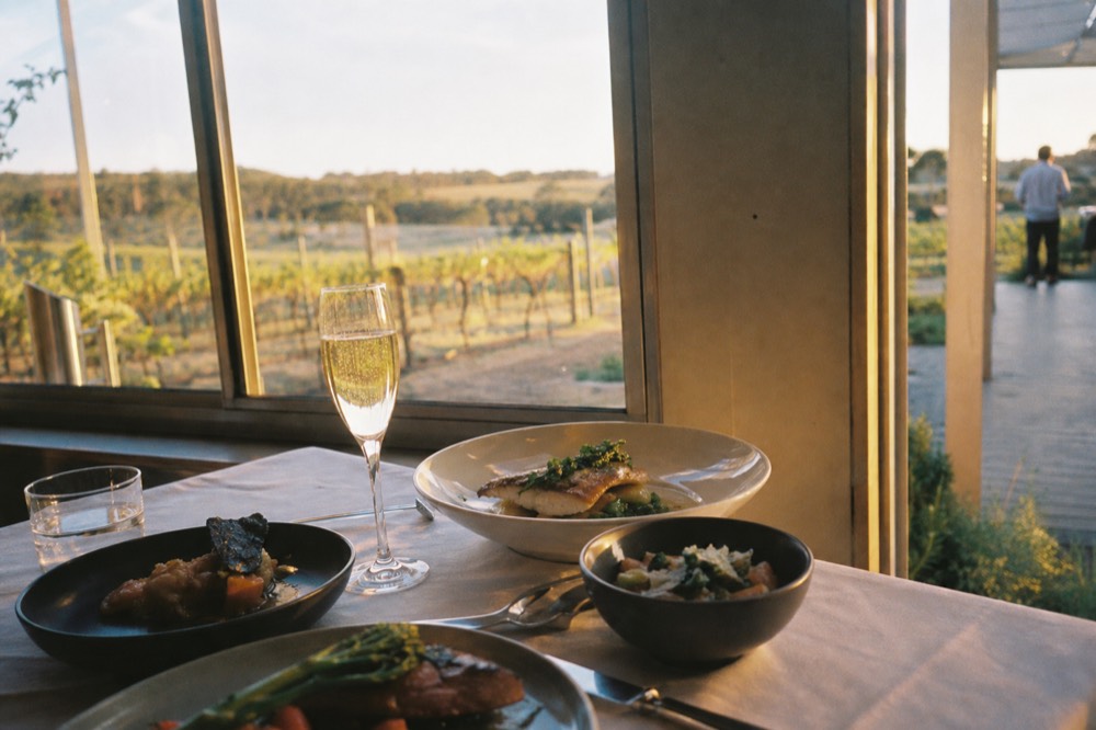 A plated multi-course meal on a white tablecloth beside a glass of sparkling wine with vineyards blurred through the window, no text, no logos, no people